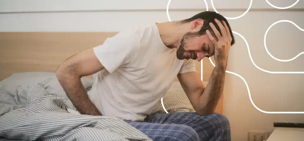Homem sentado na cama com expressão de dor, segurando a cabeça e a lombar, representando sintomas comuns da fibromialgia como dor crônica e fadiga ao acordar.